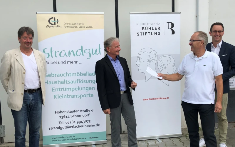 Vier M&auml;nner stehen vor zwei Infobannern mit den Aufschriften "Strandgut" und "B&uuml;hler Stiftung". Der Mann im Vordergrund auf der rechten Seite h&auml;lt ein Papier in der Hand.