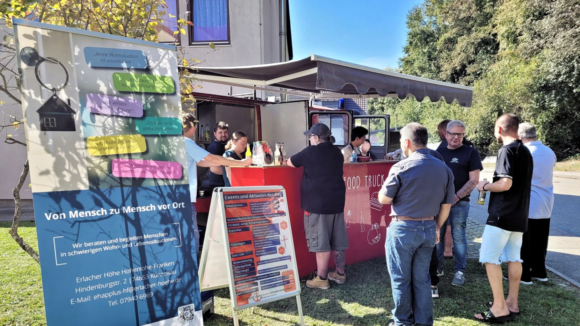 Informationsstand im Freien mit Bannern und Besuchern, die an einem Verkaufsstand stehen, vor einem Geb&auml;ude auf einer Wiese an einem sonnigen Tag.
