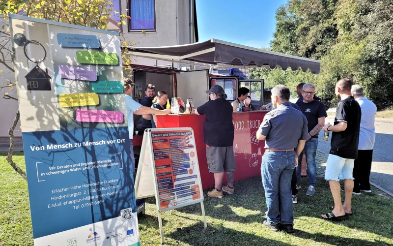 Informationsstand im Freien mit Bannern und Besuchern, die an einem Verkaufsstand stehen, vor einem Geb&auml;ude auf einer Wiese an einem sonnigen Tag.