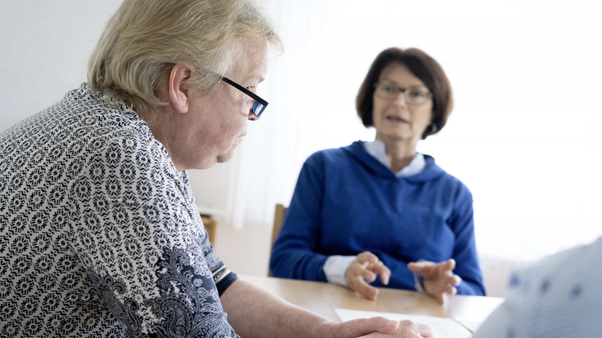 Eine Frau mit gemustertem Oberteil und Brille bekleidet sitzt im Vordergrund an einem Tisch und h&ouml;rt einer weiteren Frau in blauem Oberteil und Brille im Hintergrund zu. Eine weitere Person sitzt am rechten Bildrand.