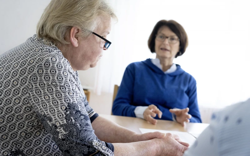 Eine Frau mit gemustertem Oberteil und Brille bekleidet sitzt im Vordergrund an einem Tisch und h&ouml;rt einer weiteren Frau in blauem Oberteil und Brille im Hintergrund zu. Eine weitere Person sitzt am rechten Bildrand.