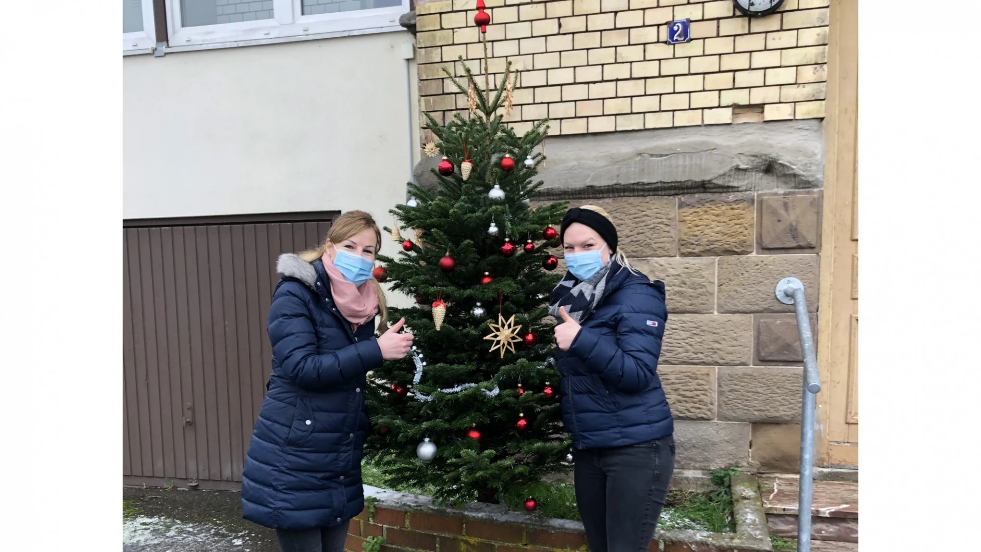 Zwei Frauen stehen mit Mund-Nasenschutz vor einem Geb&auml;ude, zwischen ihnen ein geschm&uuml;ckter Tannenbaum. Sie zeigen mit dem Daumen nach oben.
