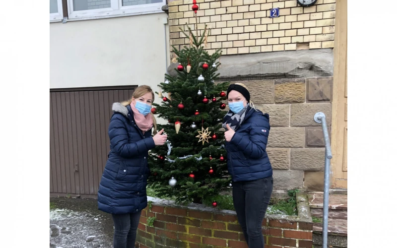 Zwei Frauen stehen mit Mund-Nasenschutz vor einem Geb&auml;ude, zwischen ihnen ein geschm&uuml;ckter Tannenbaum. Sie zeigen mit dem Daumen nach oben.