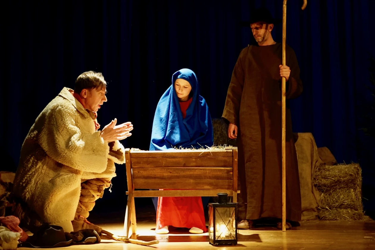 Eine Szene mit drei Figuren: Eine Frau mit einem blauen Kopftuch steht hinter einer Krippe, ein Mann in beigen Mantel in der Hocke ist daneben und eine dritte Person im braunen Umhang steht mit einem Hirtenstab daneben. Im Vordergrund ist eine Laterne zu sehen.
