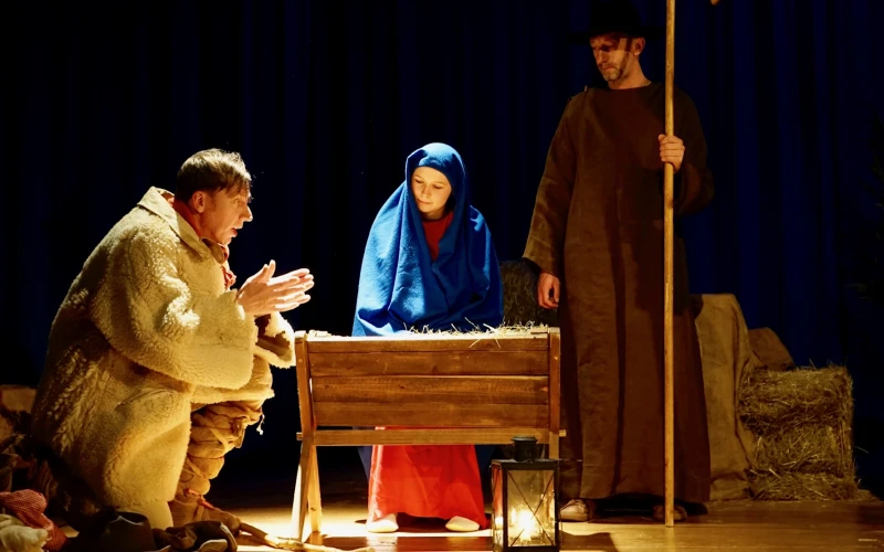 Eine Szene mit drei Figuren: Eine Frau mit einem blauen Kopftuch steht hinter einer Krippe, ein Mann in beigen Mantel in der Hocke ist daneben und eine dritte Person im braunen Umhang steht mit einem Hirtenstab daneben. Im Vordergrund ist eine Laterne zu sehen.