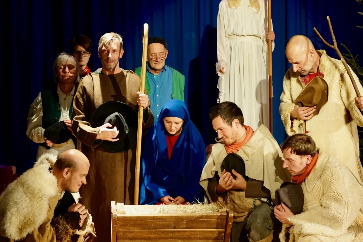 Szenische Darstellung der Weihnachtsgeschichte mit Figuren in historischen Kost&uuml;men, die um eine Krippe mit dem Jesuskind versammelt sind, vor einem dunkelblauen Hintergrund und einem goldenen Stern &uuml;ber der Szene.