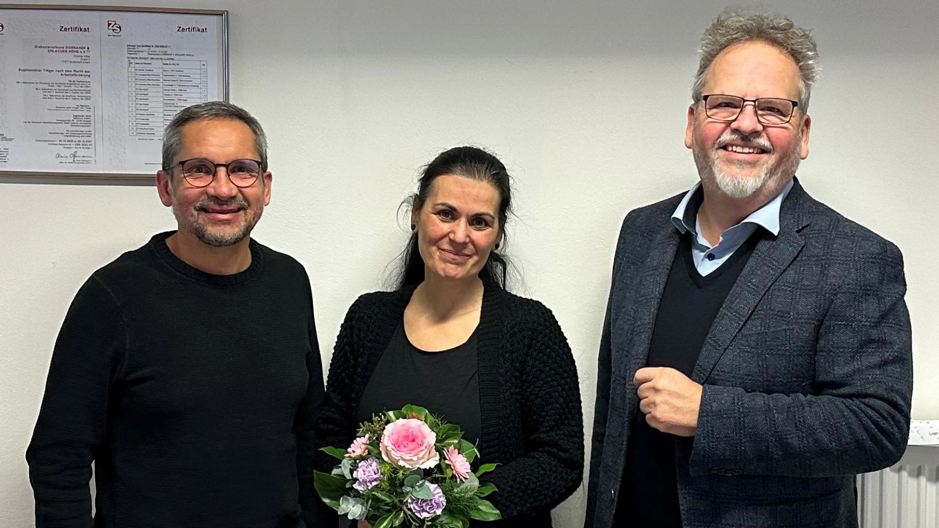 Eine Frau in dunkler Kleidung mit einem Blumenstrau&szlig; steht zwischen zwei M&auml;nnern in dunkler Kleidung. Alle drei l&auml;cheln freundlich in die Kamera.