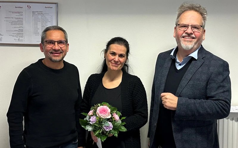 Eine Frau in dunkler Kleidung mit einem Blumenstrau&szlig; steht zwischen zwei M&auml;nnern in dunkler Kleidung. Alle drei l&auml;cheln freundlich in die Kamera.