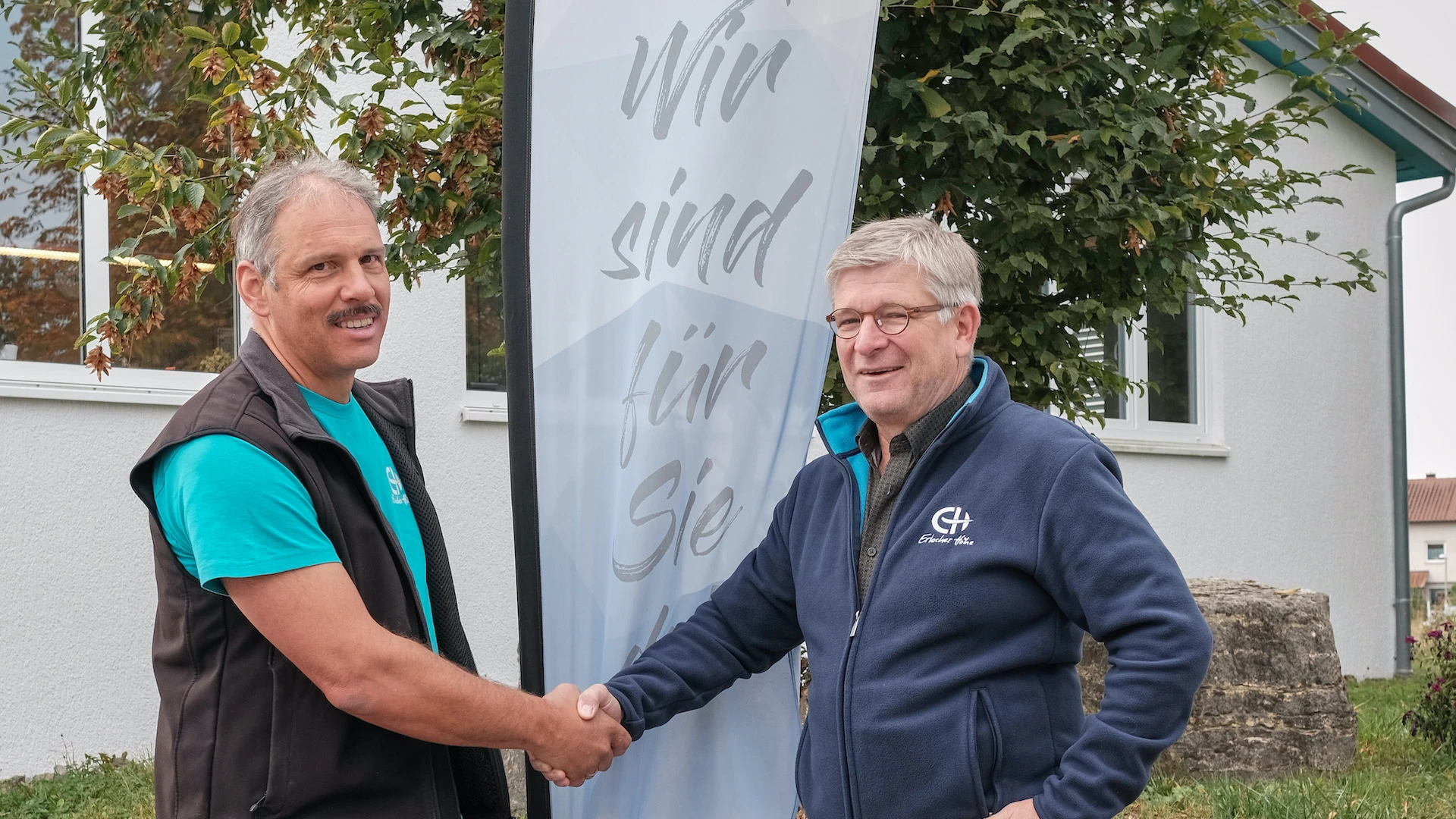 Zwei M&auml;nner geben sich die Hand, zwischen ihnen steht eine Fahne mit der Aufschrift: "Wir sind f&uuml;r Sie da". Im Hintergrund ist ein Geb&auml;ude und ein Baum zu sehen.