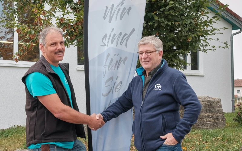 Zwei M&auml;nner geben sich die Hand, zwischen ihnen steht eine Fahne mit der Aufschrift: "Wir sind f&uuml;r Sie da". Im Hintergrund ist ein Geb&auml;ude und ein Baum zu sehen.