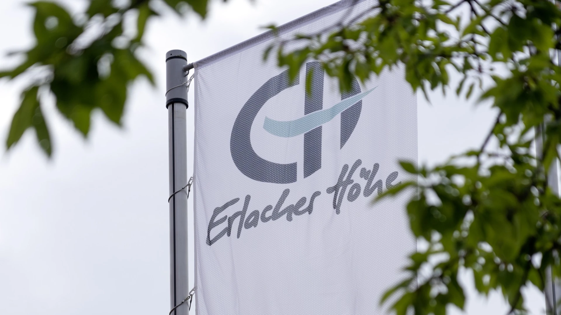 Wei&szlig;e Fahne mit dem Logo und Schriftzug der Erlacher H&ouml;he, vor gr&uuml;nen Bl&auml;ttern und einem bew&ouml;lkten Himmel. Die Flagge zeigt die Webadresse www.erlacher-hoehe.de und einen Kreis mit dem Slogan "Menschen. Leben. W&uuml;rde".