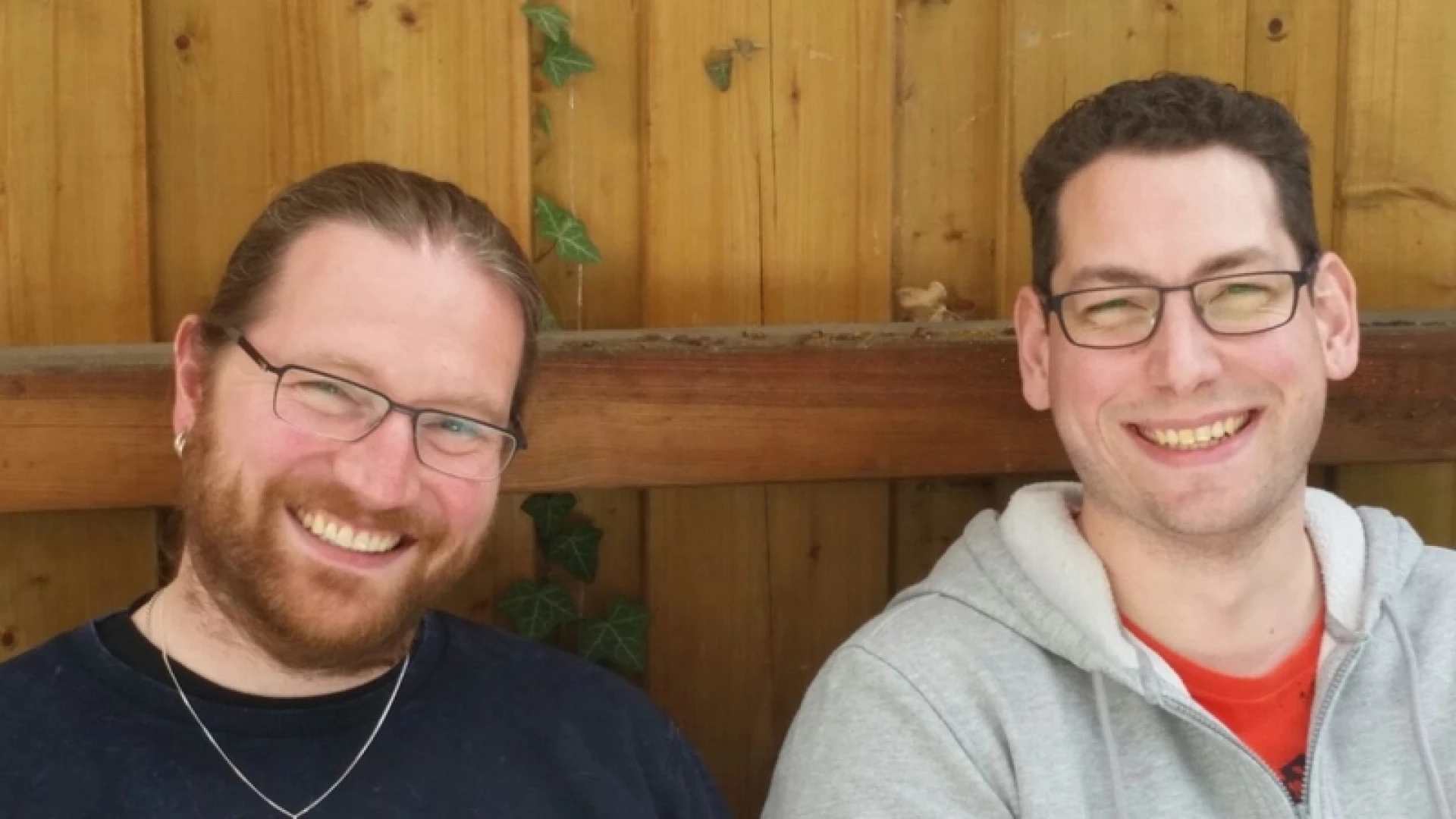 Zwei l&auml;chelnde M&auml;nner mit Brille vor einer Holzwand. Der Mann auf der linken Seite tr&auml;gt ein dunkles Shirt, eine Halskette und hat einen Bart. Der Mann rechts ist in einem grauen Kapuzenpullover gekleidet.