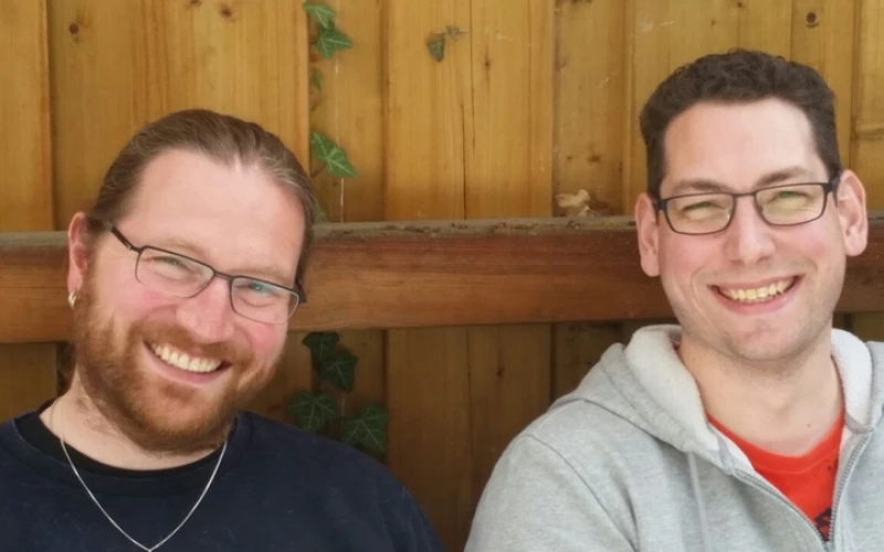 Zwei l&auml;chelnde M&auml;nner mit Brille vor einer Holzwand. Der Mann auf der linken Seite tr&auml;gt ein dunkles Shirt, eine Halskette und hat einen Bart. Der Mann rechts ist in einem grauen Kapuzenpullover gekleidet.