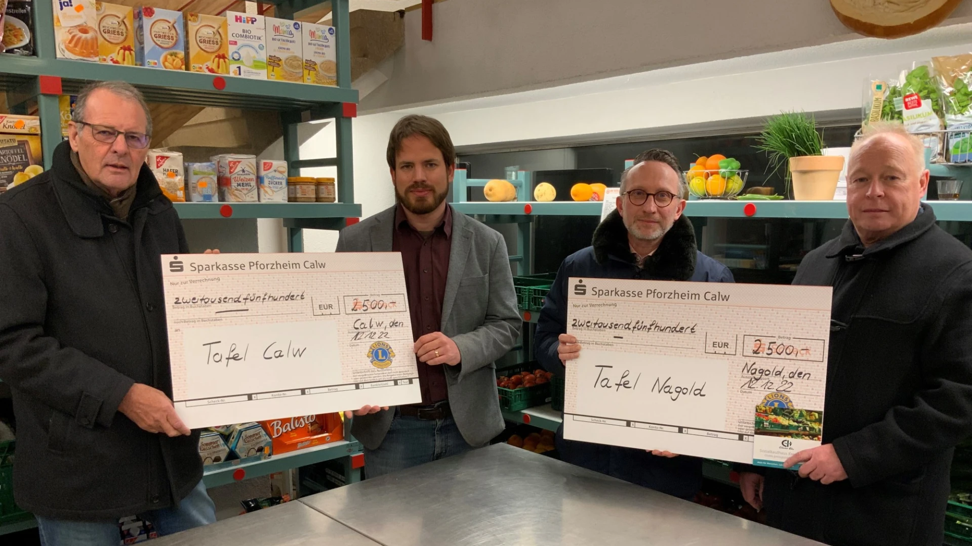Vier M&auml;nner stehen vor einer Theke in einem Lebensmittelladen in die Kamera blickend. Sie halten zwei Spendenschecks.
