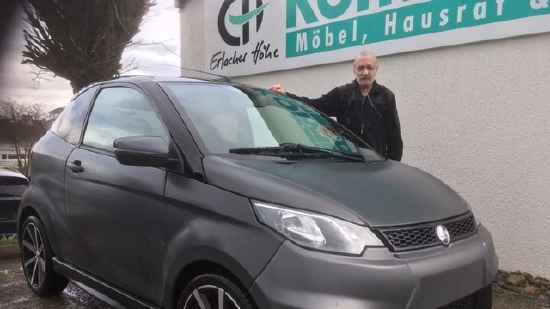 Ein Mann steht neben einem kleinen, grauen Wagen und legt die rechte Hand aufs Dach. Das Fahrzeug ist geparkt und im Hintergrund befindet sich eine wei&szlig;e Geb&auml;udewand mit dem Logo und Schriftzug der Erlacher H&ouml;he.