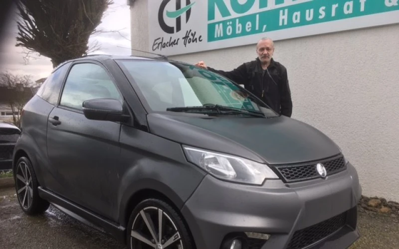 Ein Mann steht neben einem kleinen, grauen Wagen und legt die rechte Hand aufs Dach. Das Fahrzeug ist geparkt und im Hintergrund befindet sich eine wei&szlig;e Geb&auml;udewand mit dem Logo und Schriftzug der Erlacher H&ouml;he.