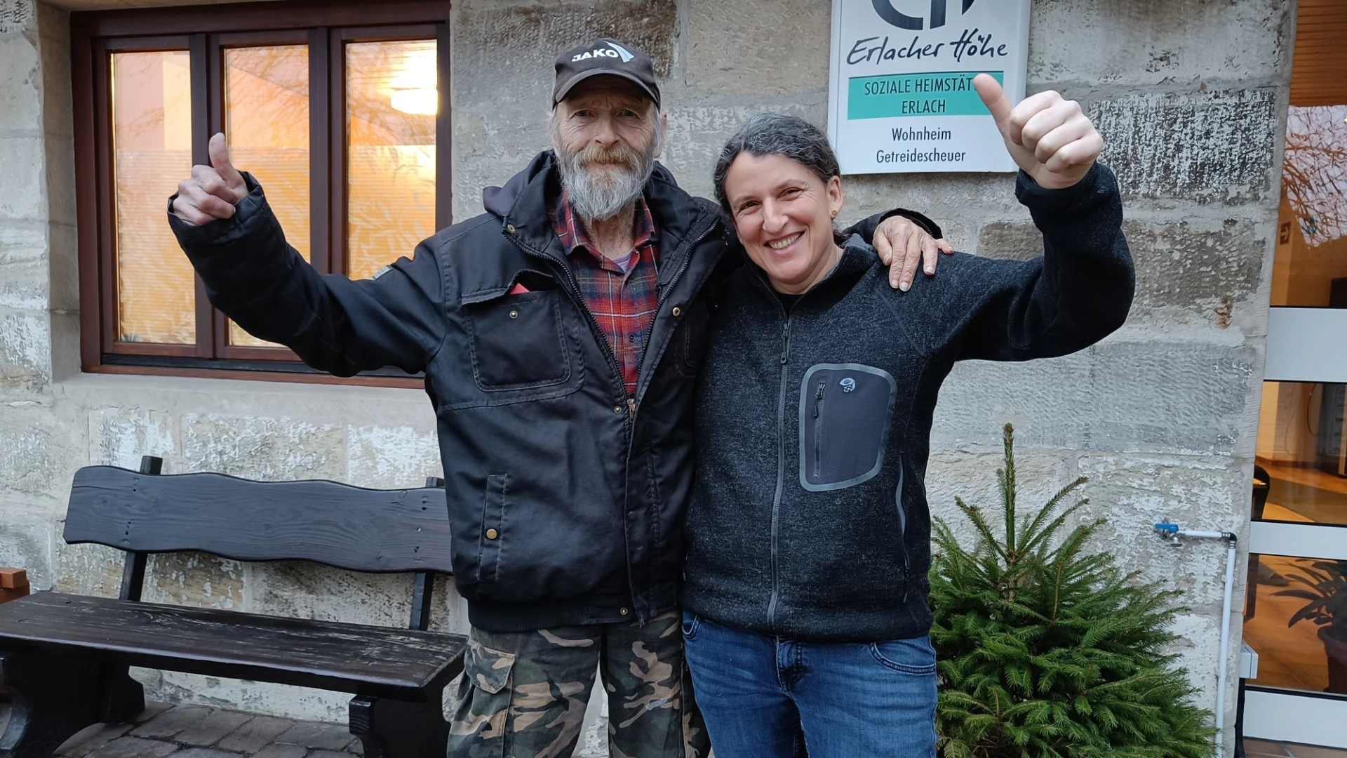 Eine Frau und ein Mann haben ihre Arme um die Schultern gelegt und zeigen jeweils den Daumen nach oben. Sie l&auml;cheln in die Kamera. Im Hintergrund h&auml;ngt an der Wand ein Schild der Erlacher H&ouml;he mit der Aufschrift "Soziale Heimst&auml;tte Erlach" und daneben steht eine Holzbank.