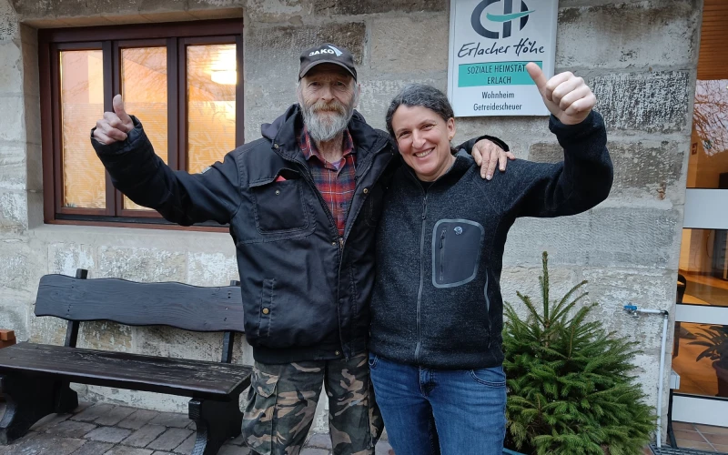 Eine Frau und ein Mann haben ihre Arme um die Schultern gelegt und zeigen jeweils den Daumen nach oben. Sie l&auml;cheln in die Kamera. Im Hintergrund h&auml;ngt an der Wand ein Schild der Erlacher H&ouml;he mit der Aufschrift "Soziale Heimst&auml;tte Erlach" und daneben steht eine Holzbank.