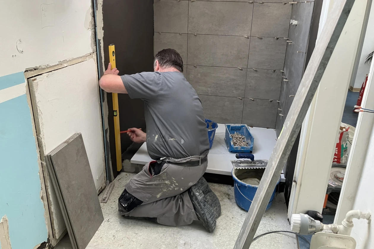 Handwerker kniet bei der Arbeit auf einer Baustelle im Badezimmer. Mit einer Wasserwaage kontrolliert er den senkrechten Wandaufbau. Im Vordergrund sind Werkzeuge und Materialien zu sehen.