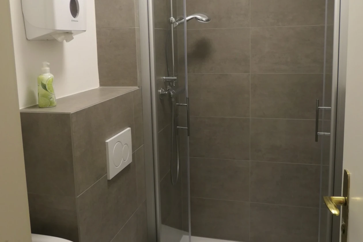 Ein renoviertes Badezimmer mit Dusche, WC und Waschbecken. Die zweifl&uuml;glige Glast&uuml;re der Dusche steht offen. Die W&auml;nde sind im Nassbereich mit dunkelgrauen Fliesen versehen, die restlichen W&auml;nde sind wei&szlig; verputzt.