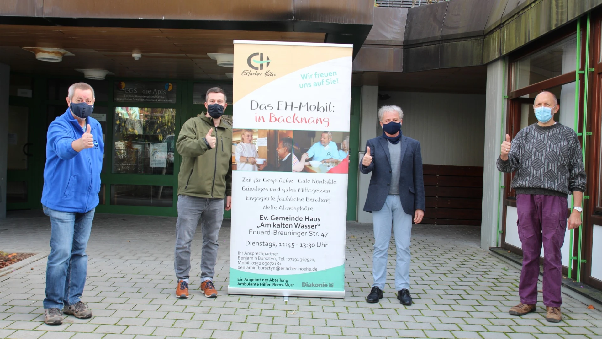 Vier Personen mit Mund-Nasenschutz stehen vor einem Geb&auml;ude auf einem Pflasterboden. Alle zeigen mit den Daumen nach oben. Zwischen ihnen befindet sich ein aufgestellter Banner mit der Aufschrift: "Das EH-Mobil in Backnang".
