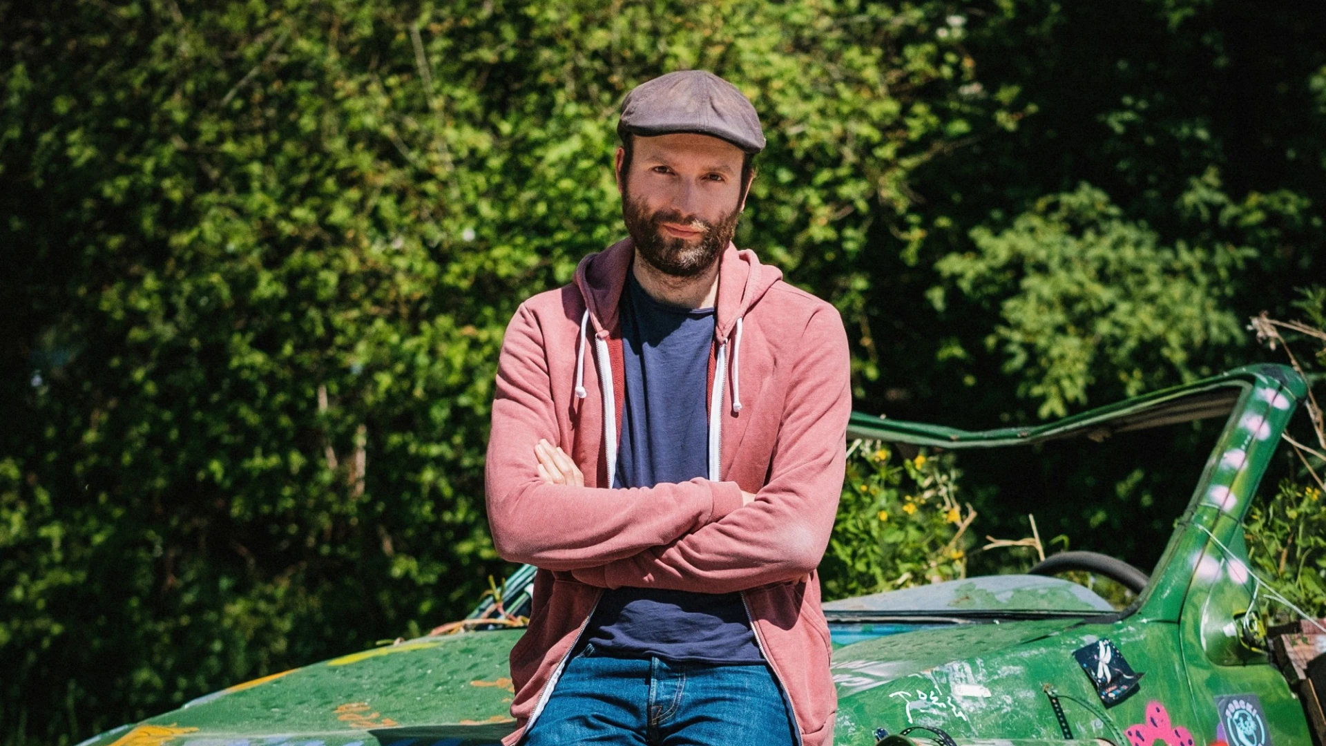 Ein junger Mann, l&auml;ssig gekleidet mit Jeans, hellroter Jacke und M&uuml;tze lehnt an einem alten gr&uuml;nen Auto. Im Hintergrund sind B&auml;ume zu sehen.