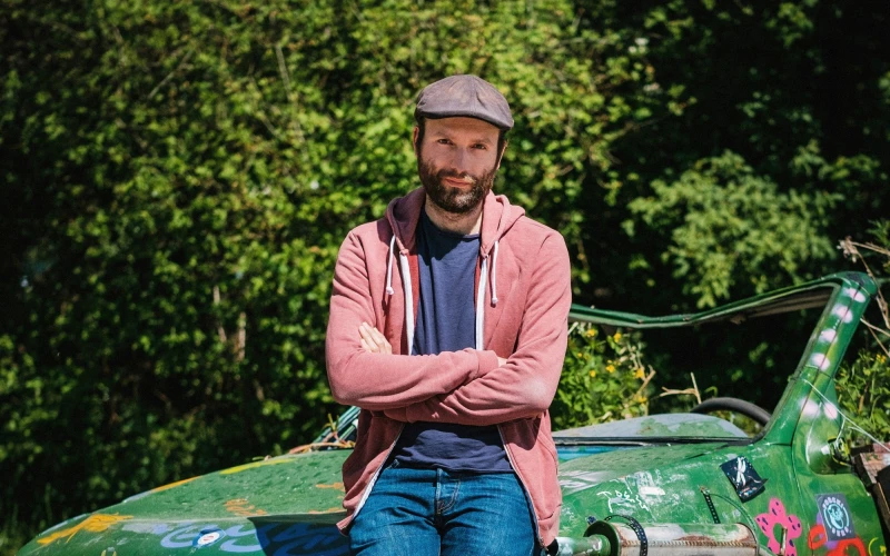 Ein junger Mann, l&auml;ssig gekleidet mit Jeans, hellroter Jacke und M&uuml;tze lehnt an einem alten gr&uuml;nen Auto. Im Hintergrund sind B&auml;ume zu sehen.