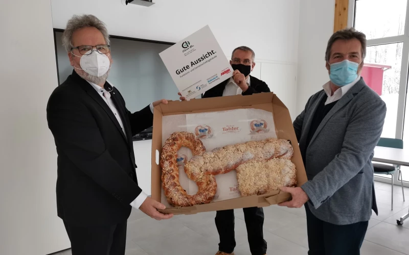 Zwei M&auml;nner halten eine gro&szlig;e Schachtel mit Geb&auml;ck in Form eines Schl&uuml;ssels in der Hand. Der Mann im Hintergrund h&auml;lt ein Schild mit der Aufschrift: "Gute Aussicht" in der Hand. Alle Personen tragen einen Mund-Nasenschutz.