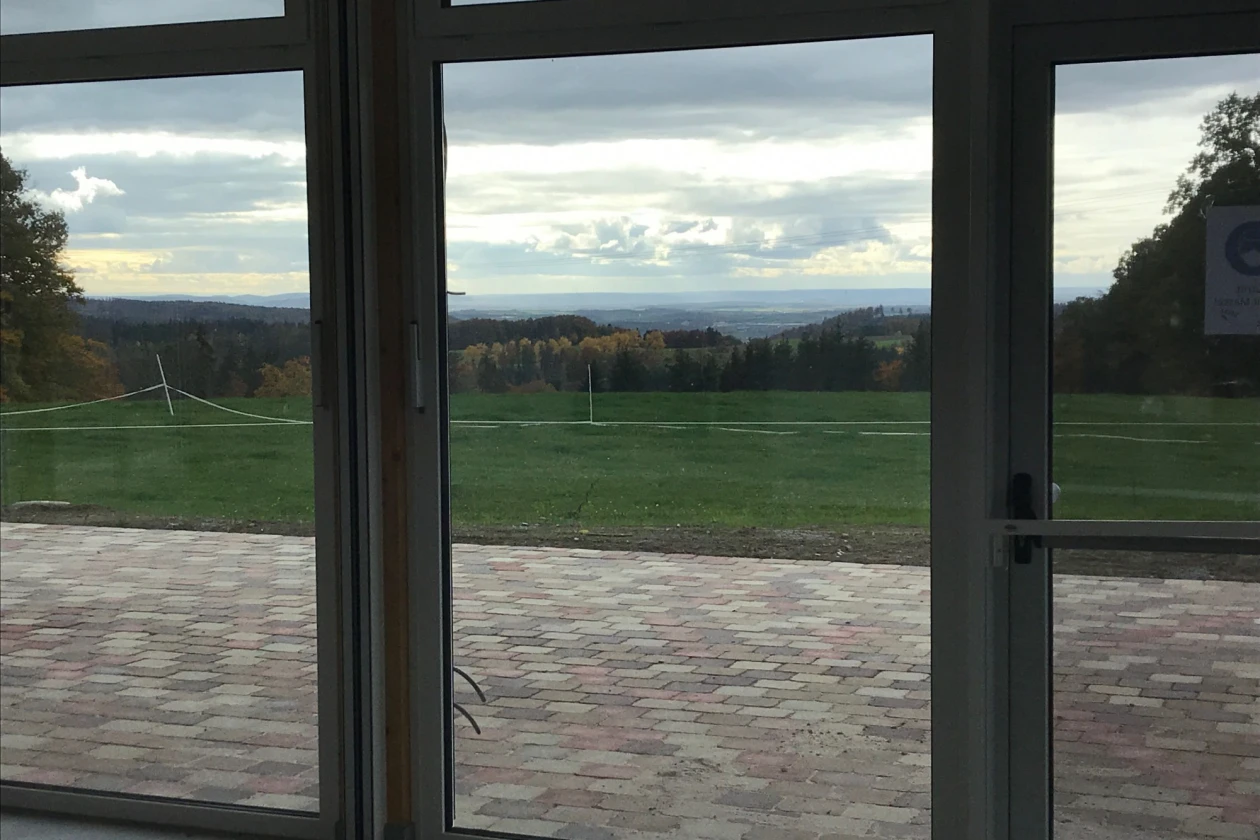 Gro&szlig;e Glasfenster bieten den Blick auf eine Terrasse und dahinter auf eine Wiese, die an ein Waldgebiet grenzt.