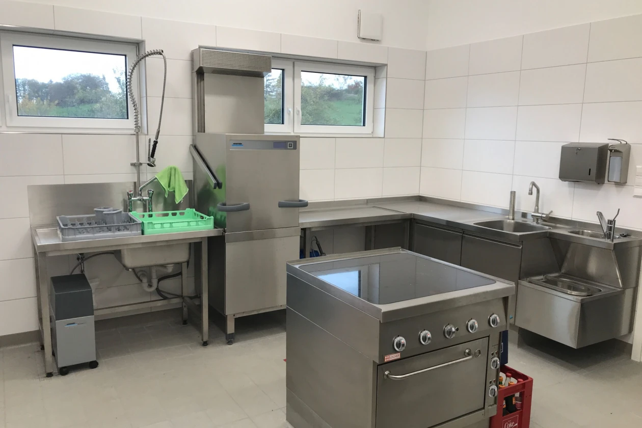 Das Bild zeigt eine leere Gro&szlig;k&uuml;che mit Edelstahlger&auml;ten und Arbeitsfl&auml;chen. Sichtbar sind ein Herd, zwei Sp&uuml;lbecken, sowie eine Ablagefl&auml;che. Die K&uuml;che verf&uuml;gt zudem &uuml;ber drei Fenster.