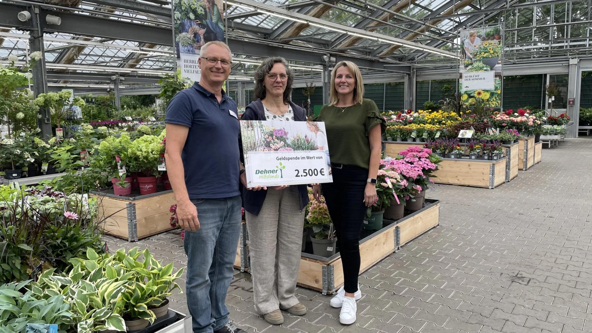 Drei Personen stehen in einer G&auml;rtnerei und halten einen Spendenscheck &uuml;ber 2.500 Euro in die Kamera