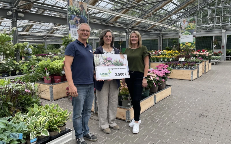 Drei Personen stehen in einer G&auml;rtnerei und halten einen Spendenscheck &uuml;ber 2.500 Euro in die Kamera