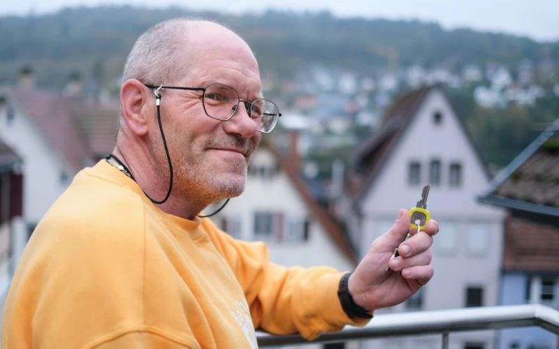 &Auml;lterer Mann mit Brille in gelbem Pullover l&auml;chelt freundlich, w&auml;hrend er auf einem Balkon mit H&auml;usern im Hintergrund steht und einen Schl&uuml;ssel in der Hand h&auml;lt.
