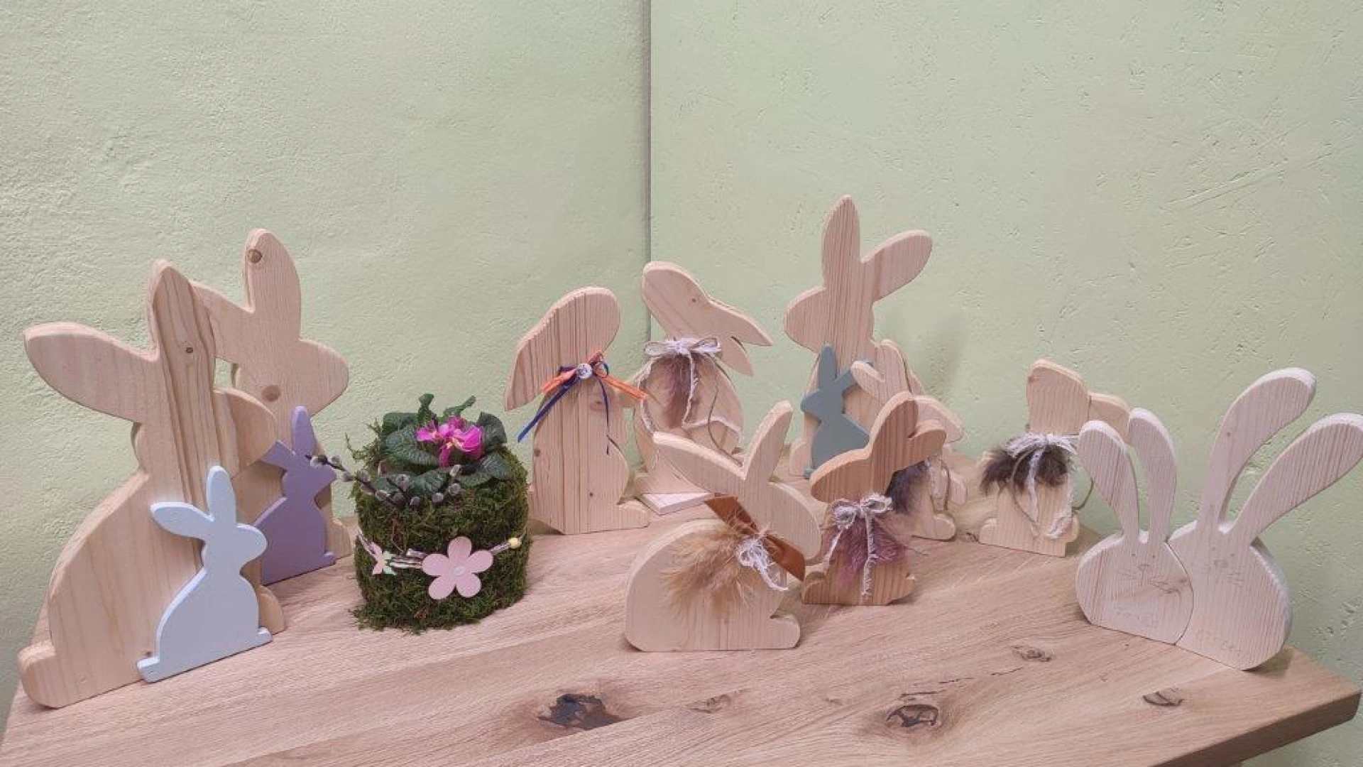 Holzhasen in verschiedenen Gr&ouml;&szlig;en, in Holz- und Pastellfarben stehen auf einem Holztisch, dazwischen ein kleiner Blumentopf mit Moos und Bl&uuml;ten. Die Hasen sind in unterschiedlichen Posen und Stellungen angeordnet, was eine s&uuml;&szlig;e, rustikale Osterdekoration ergibt.