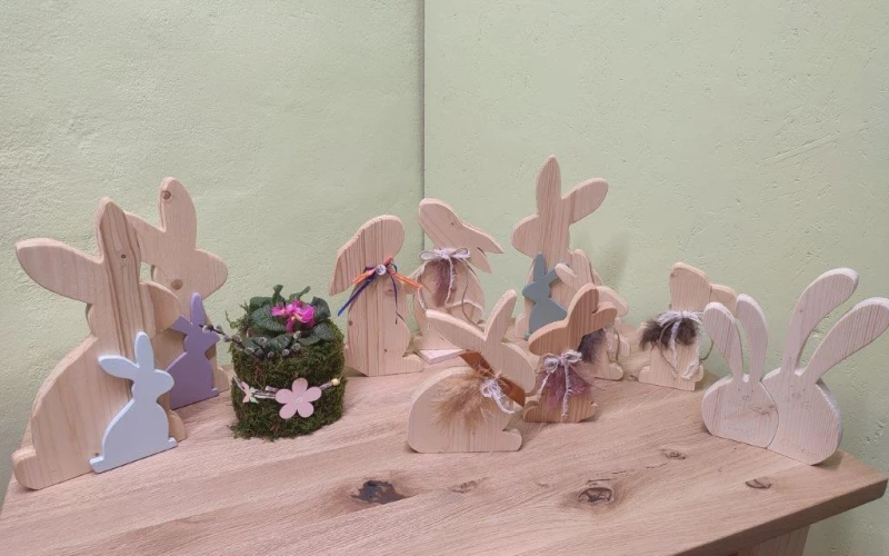 Holzhasen in verschiedenen Gr&ouml;&szlig;en, in Holz- und Pastellfarben stehen auf einem Holztisch, dazwischen ein kleiner Blumentopf mit Moos und Bl&uuml;ten. Die Hasen sind in unterschiedlichen Posen und Stellungen angeordnet, was eine s&uuml;&szlig;e, rustikale Osterdekoration ergibt.