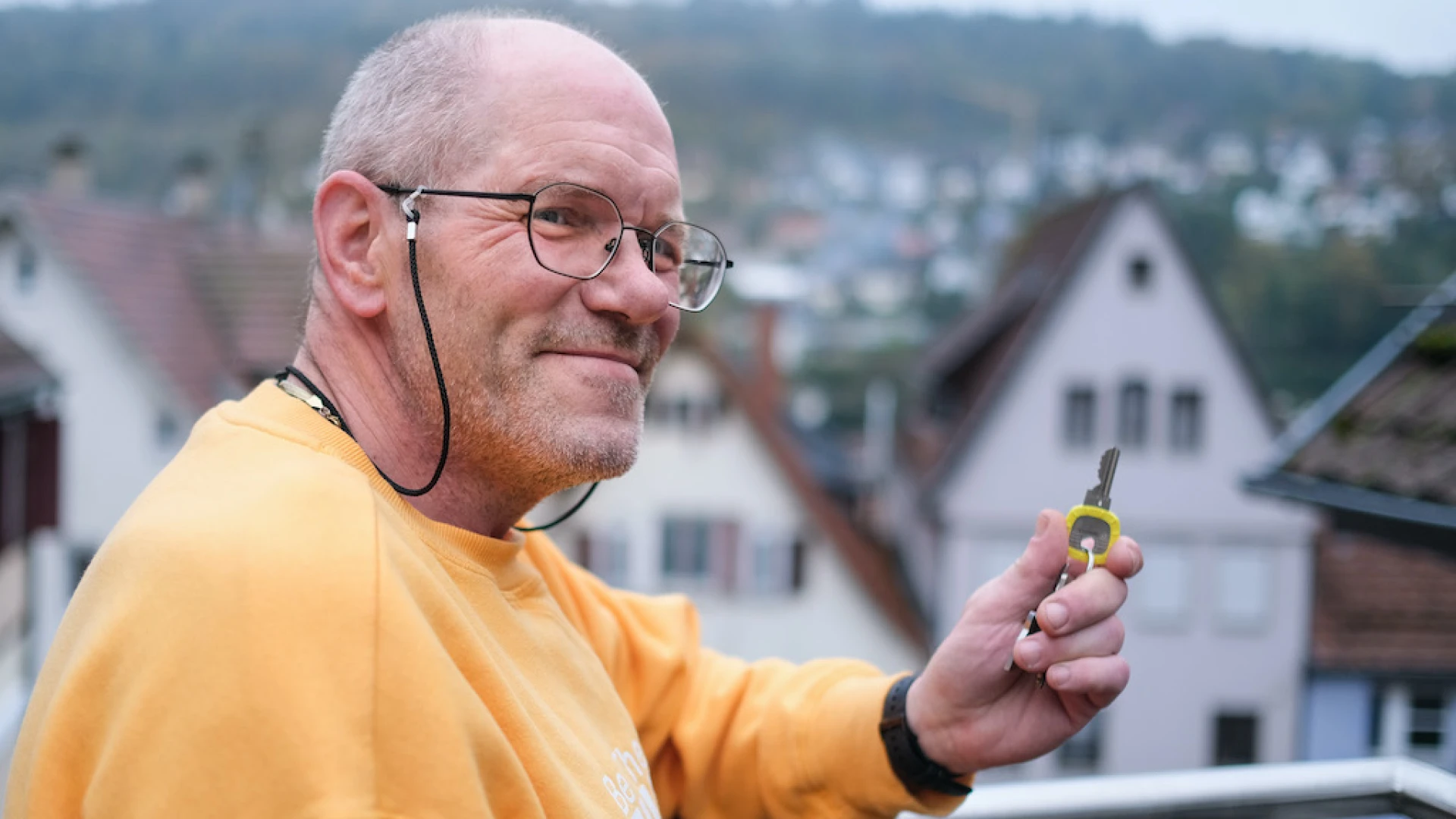 &Auml;lterer Mann mit Brille in gelbem Pullover l&auml;chelt freundlich, w&auml;hrend er auf einem Balkon mit H&auml;usern im Hintergrund steht und einen Schl&uuml;ssel in der Hand h&auml;lt.