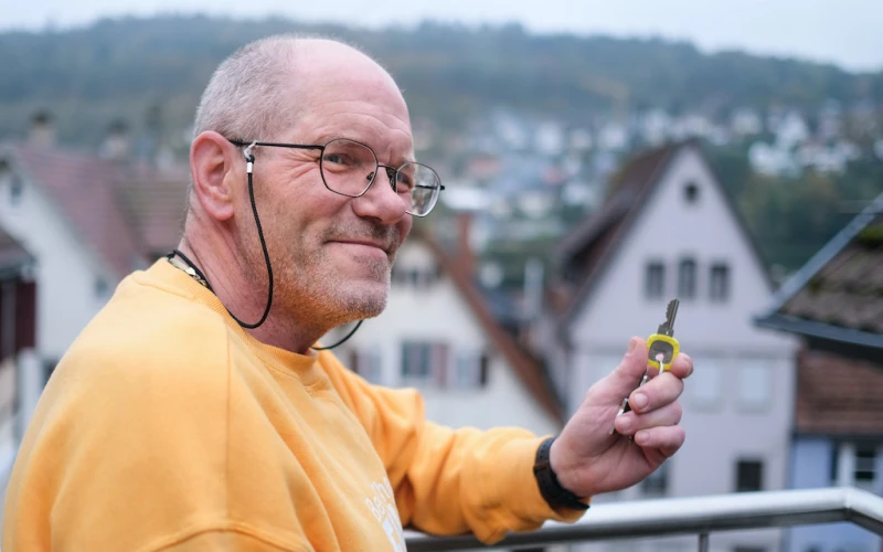 &Auml;lterer Mann mit Brille in gelbem Pullover l&auml;chelt freundlich, w&auml;hrend er auf einem Balkon mit H&auml;usern im Hintergrund steht und einen Schl&uuml;ssel in der Hand h&auml;lt.