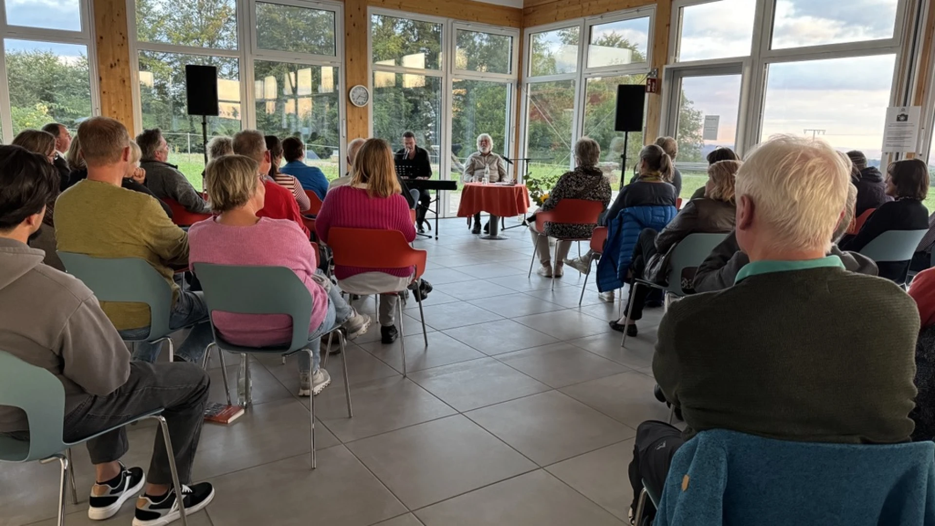 Menschen sitzen auf St&uuml;hlen in einem hellen lichtdurchfluteten Raum mit Holzbalken und gro&szlig;en Fenstern und h&ouml;ren einem Vortragenden an einem Tisch zu. Daneben sitzt ein Mann an einem E-Piano.