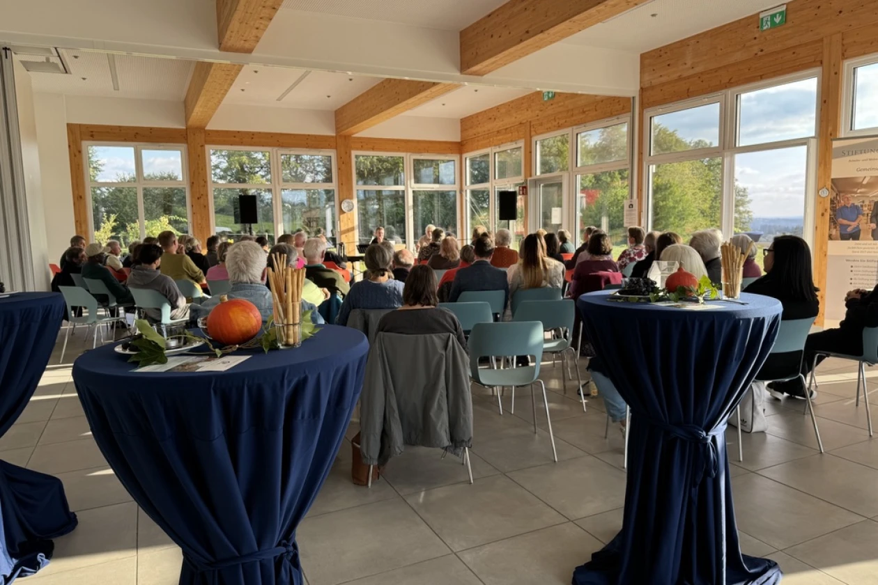 Menschen sitzen auf St&uuml;hlen in einem hellen lichtdurchfluteten Raum Mit Holzbalken und gro&szlig;en Fenstern.mit bodentiefen Fensterfronten und h&ouml;ren einem Vortragenden an einem Tisch zu. Im Vordergrund sind Stehtische mit herbstlicher Deko zu sehen.