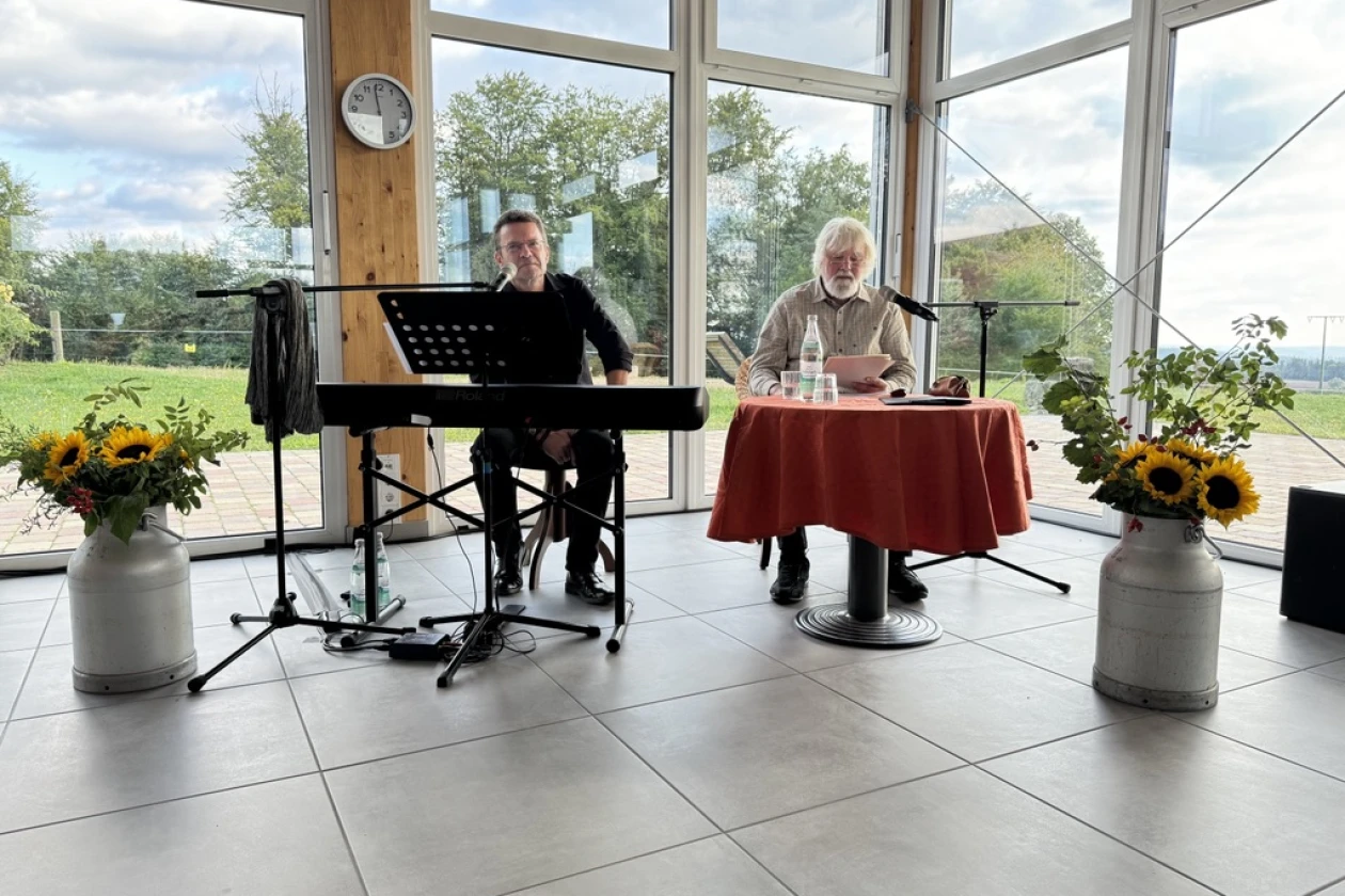 Zwei M&auml;nner im Raum mit gro&szlig;en Fenstern und Blick ins Gr&uuml;ne, einer sitzt an einem Keyboard, der andere an einem rot eingedeckten Tisch. Sonnenblumen in Milchkannen schm&uuml;cken den Raum.