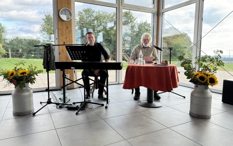 Zwei M&auml;nner im Raum mit gro&szlig;en Fenstern und Blick ins Gr&uuml;ne, einer sitzt an einem Keyboard, der andere an einem rot eingedeckten Tisch. Sonnenblumen in Milchkannen schm&uuml;cken den Raum.