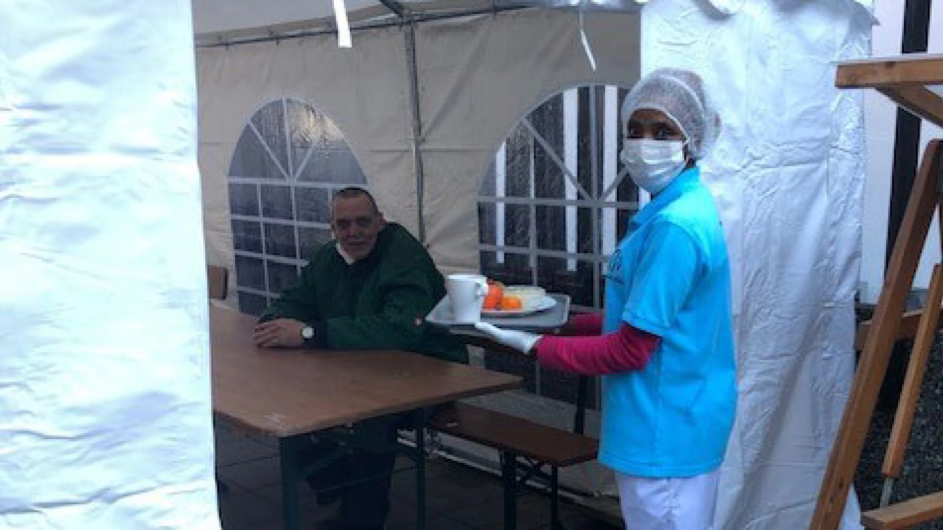 Eine Frau in Schutzkleidung mit Maske und Haube, tr&auml;gt ein Tablett mit einer Mahlzeit und einem Getr&auml;nk. Im Hintergrund sitzt ein Mann an einem Holztisch in einem wei&szlig;en Pavillonzelt mit Fenstereinsatz.