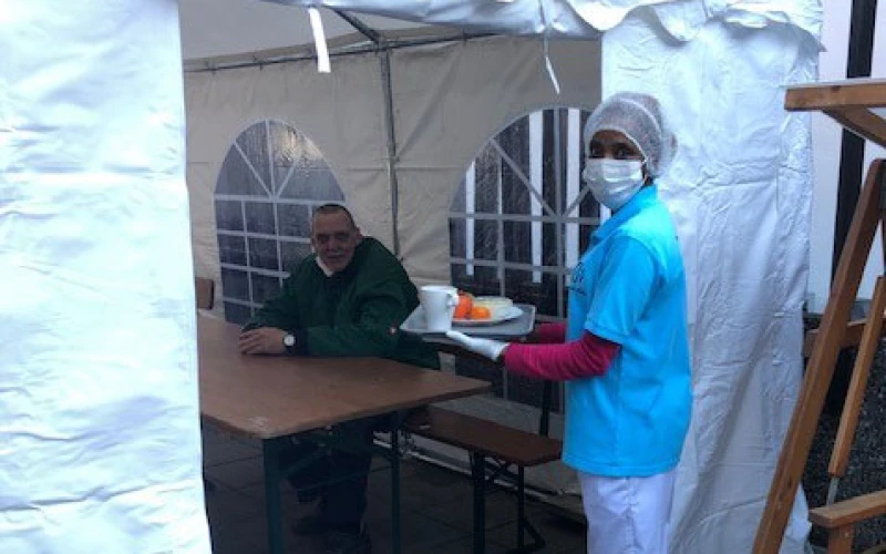 Eine Frau in Schutzkleidung mit Maske und Haube, tr&auml;gt ein Tablett mit einer Mahlzeit und einem Getr&auml;nk. Im Hintergrund sitzt ein Mann an einem Holztisch in einem wei&szlig;en Pavillonzelt mit Fenstereinsatz.
