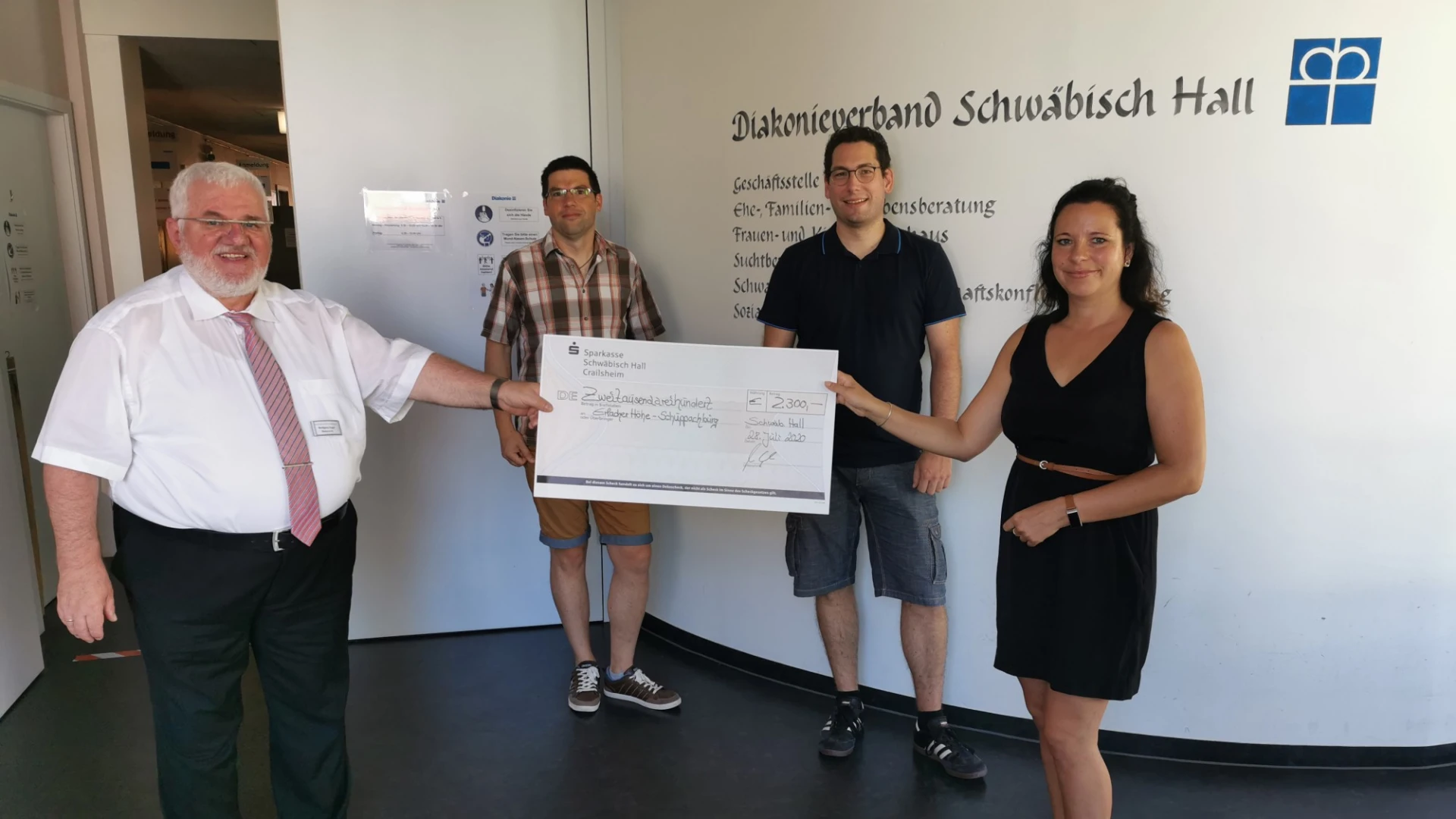 Vier Personen stehen in einem Raum vor einer Wand mit der Aufschrift: "Diakonieverbund Schw&auml;bisch Hall". Zwei Personen, die im Vordergrund stehen halten gemeinsam einen Scheck in der Hand.