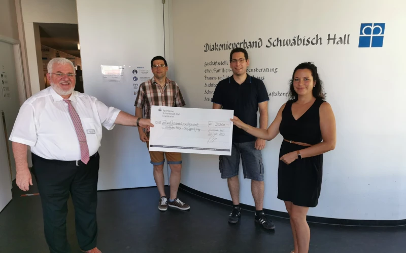Vier Personen stehen in einem Raum vor einer Wand mit der Aufschrift: "Diakonieverbund Schw&auml;bisch Hall". Zwei Personen, die im Vordergrund stehen halten gemeinsam einen Scheck in der Hand.