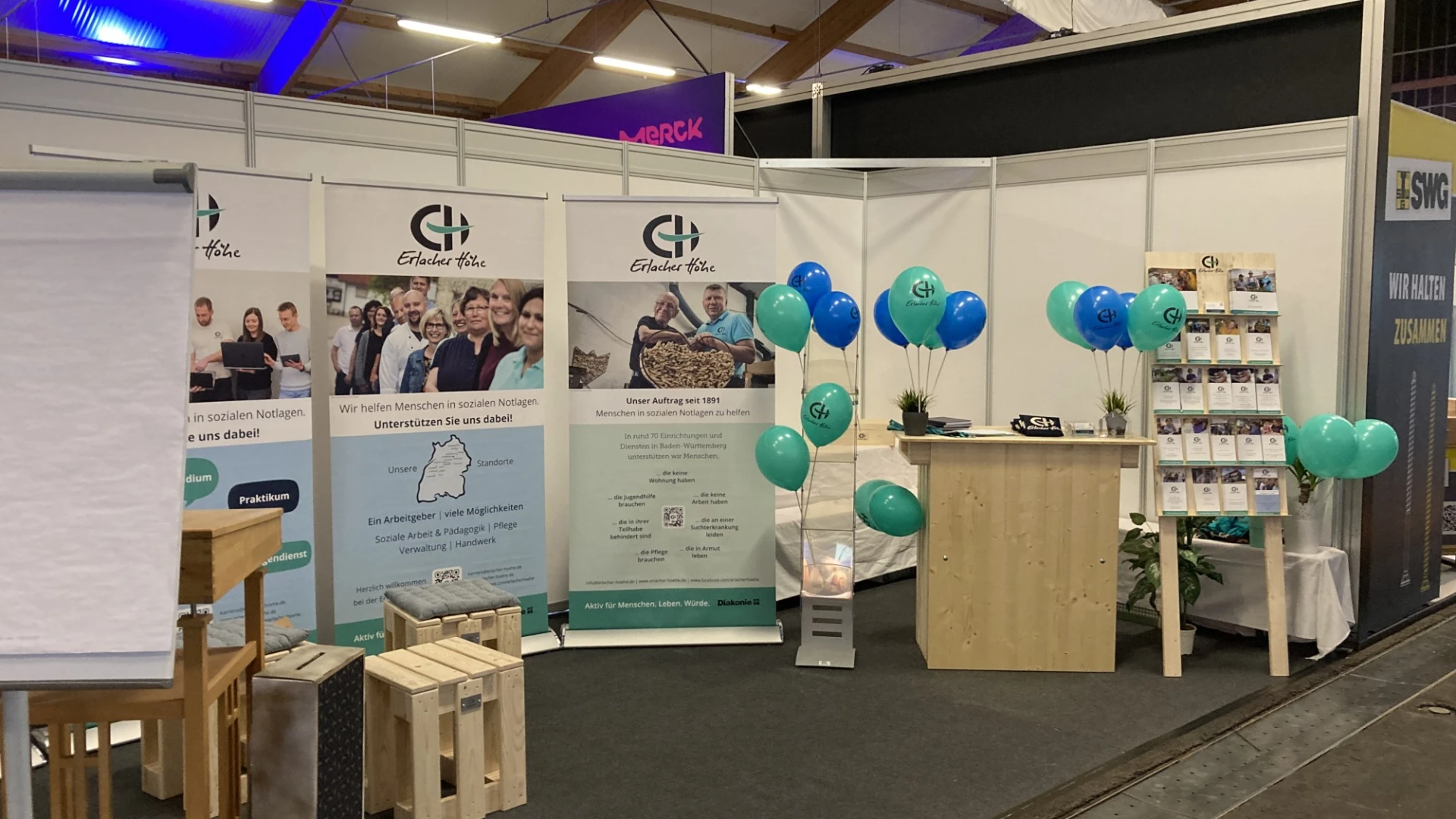 Messestand der Erlacher H&ouml;he mit wei&szlig;en Trennw&auml;nden, Bannern und Informationstafeln. Blau-gr&uuml;ne Luftballons dekorieren den Stand, der auf einer Messe oder Veranstaltung pr&auml;sentiert wird. Holzkisten und ein Informationstresen vervollst&auml;ndigen den professionell gestalteten Ausstellungsbereich.
