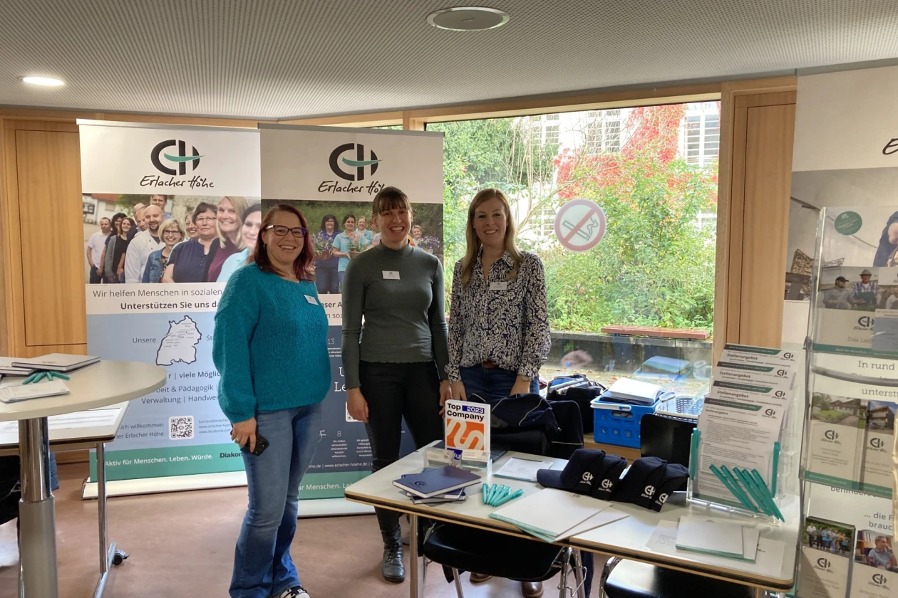 Drei Frauen stehen an einem Messestand des Erlacher H&ouml;he mit Rollups, Informationsmaterialien und Ausstellungstisch, vor einem Fenster mit Blick ins Gr&uuml;ne.