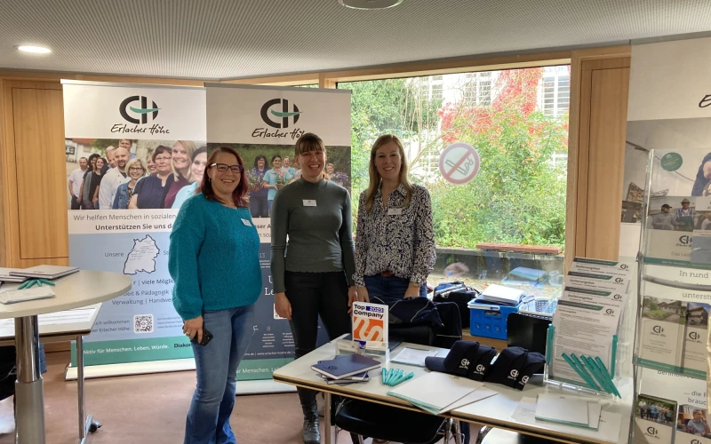 Drei Frauen stehen an einem Messestand des Erlacher H&ouml;he mit Rollups, Informationsmaterialien und Ausstellungstisch, vor einem Fenster mit Blick ins Gr&uuml;ne.