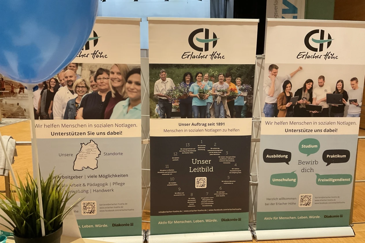 Drei Roll-ups der Erlacher H&ouml;he stehen nebeneinander, die verschiedene Aspekte der Organisation und Arbeit pr&auml;sentieren. Im Vordergrund ist ein blauer Luftballon und eine Topfpflanze zu sehen.