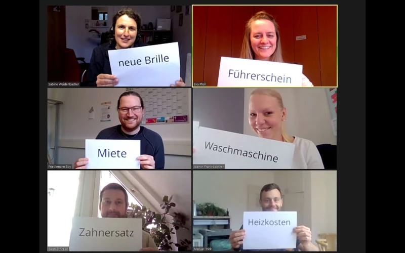 Sechs Personen in einer Videokonferenz. Sie halten Schilder mit einem Worten "neue Brille", "F&uuml;hrerschein", "Miete", "Waschmaschine", "Zahnersatz" und "Heizkosten" in die Kamera und l&auml;cheln dabei.